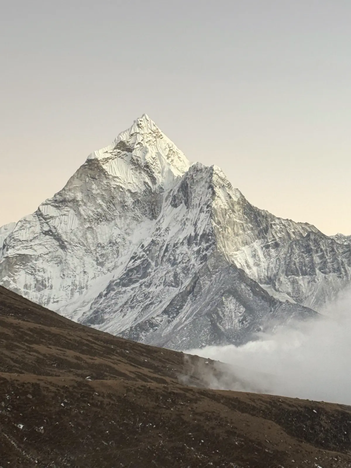 A snow-covered Himalayan peak rising above mist and brown moraine in the first light of morning.